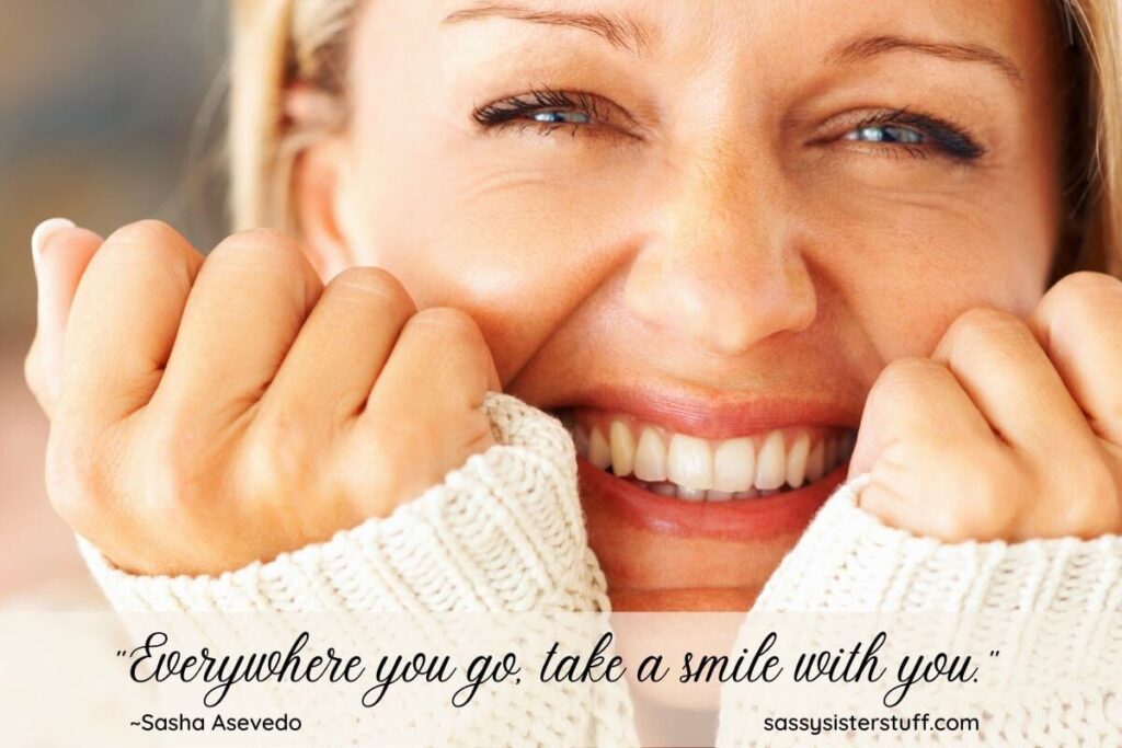 a close up of a happy woman smiling big with her chin resting on her hands wearing a cream sweater and a smile quote