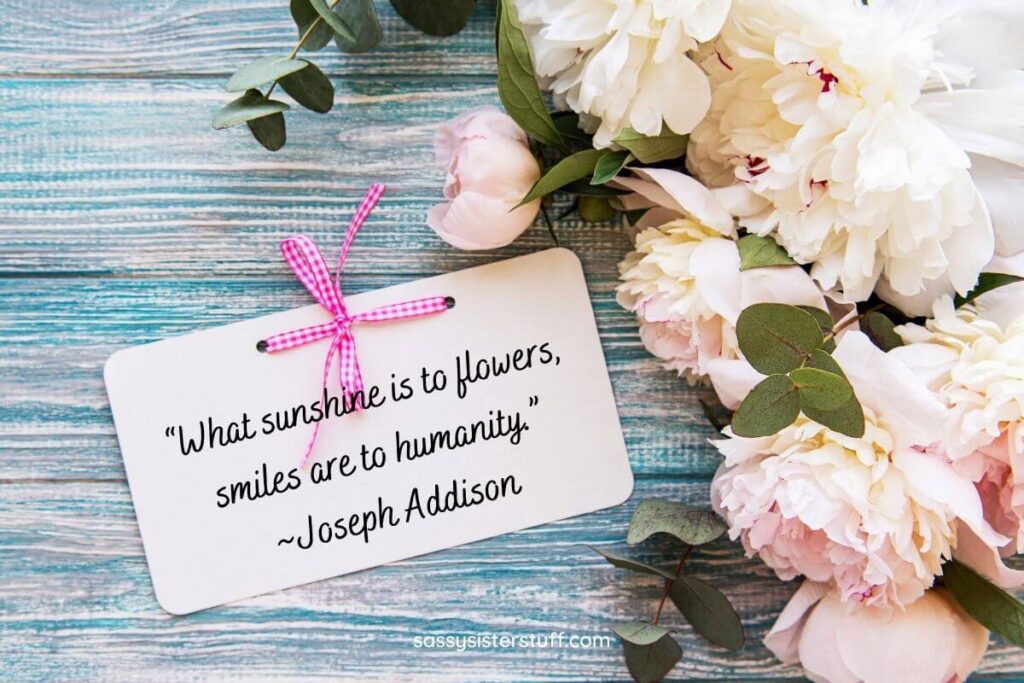 white and pink flowers laying on a wooden teal table with a tag that says what sunshine is to flowers smile are to humanity