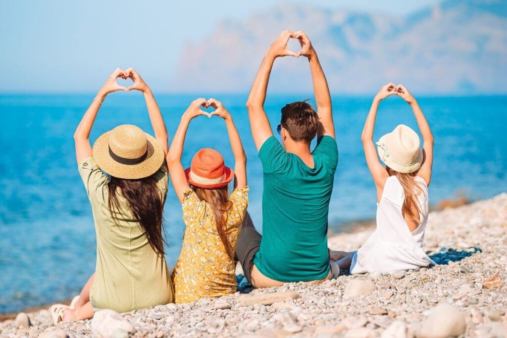 family of four with a dad mom and two young daughters sit on a rocky beach with their hands in the air making heart signs against a beautiful blue sky