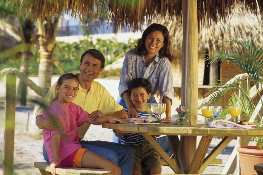 family of four includes a mom dad young boy and young girl sit at a table on a tropical island where they have taken a vacation during the school year