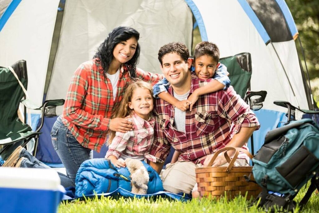 family of four with a mom dad young boy and young girl sit together outside in front of their tent on a camping trip vacation during the school year