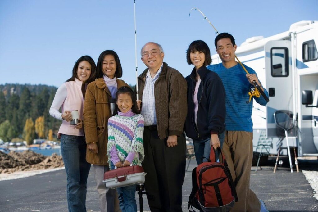 four generations of family on a vacation during the school year stand outside their RV with fishing poles in front of a lake and a beautiful blue sky