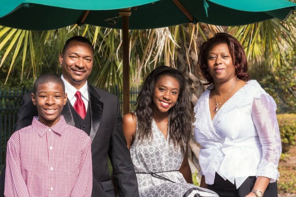 family of four with mom dad daughter and son dressed up for a wedding and posing together happily under an outdoor tent in front of palm trees