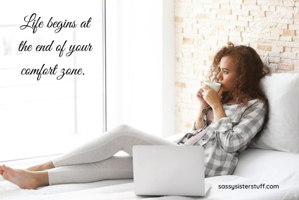 a young woman relaxes in the sunshine on a bed next to a large window with her laptop next to her as she drinks a cup of coffee and a personal growth quote say life begins at the end of your comfort zone