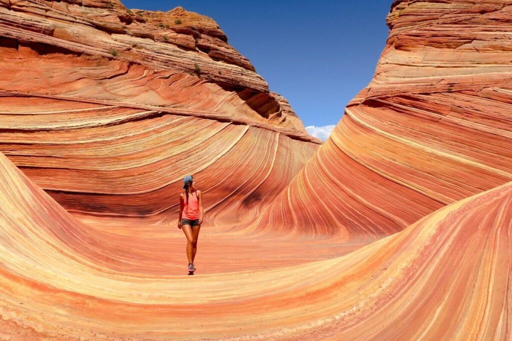 a woman in a baseball cap and summer clothes walks casually at the bottom of a very dramatic deep geological formation of swirling orange red and brown rocks against a beautiful blue sky