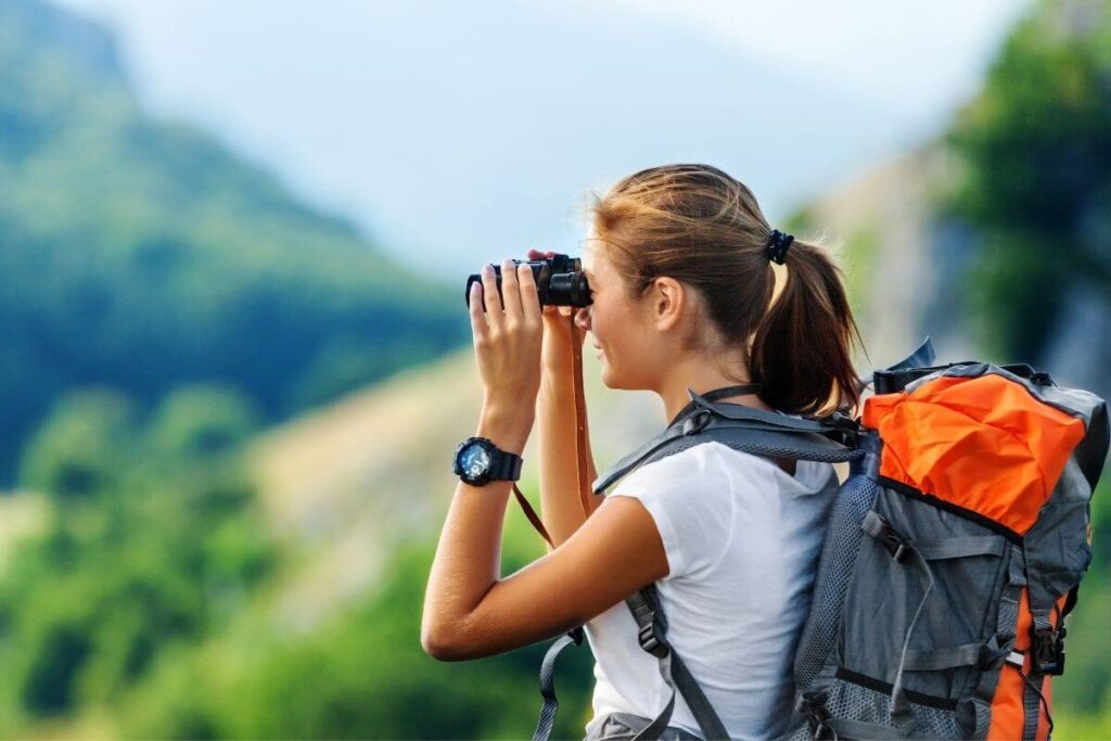 a young woman wearing a hiking backpack and a white tee shirt stands in the tree and grass covered mountains peacefully looking out over a valley with binoculars