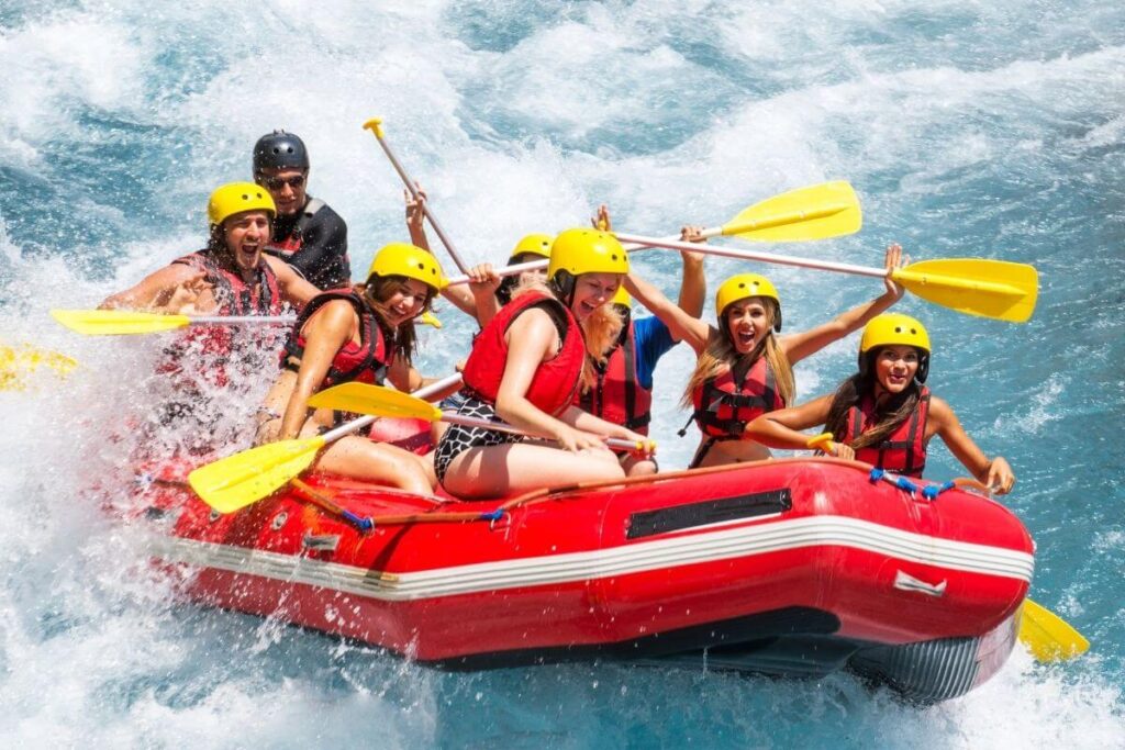 8 young men and women wearing life vests and helmets sit in a large red raft as they navigate through white water rapids