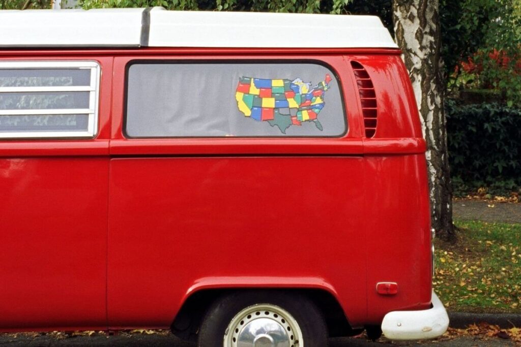 an old red volkswagen van with a map of the united states on the window sits on a curb on a tree lined park street