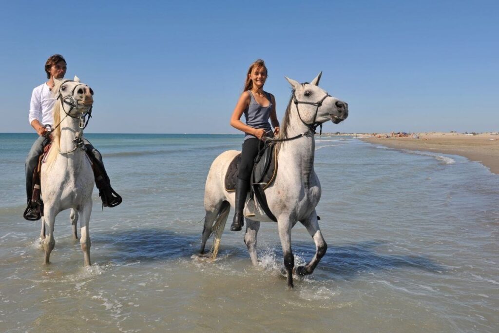 a man and a woman ride white horses along a beach in the shallow waves against a clear blue skies