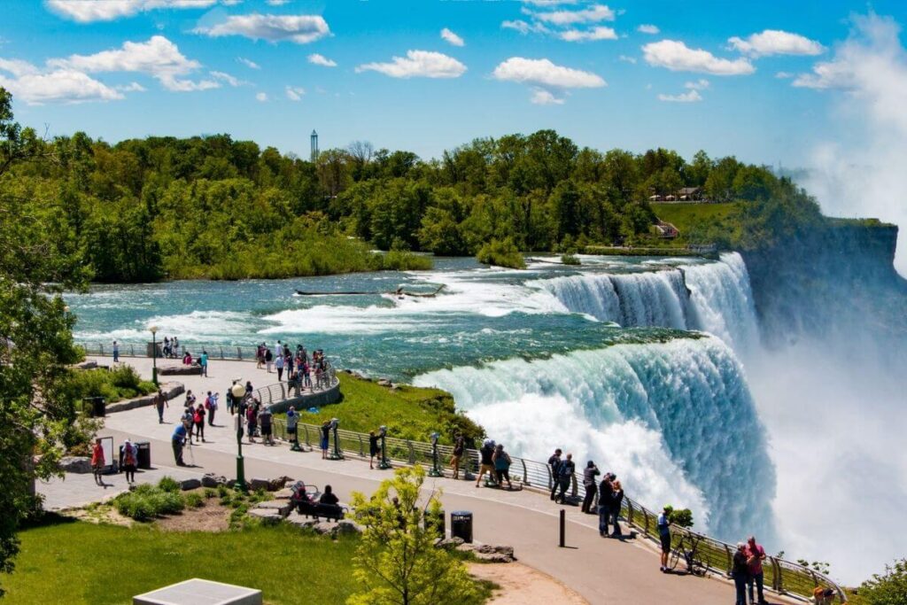 an image of the American side of the Niagara Falls with a long winding sidewalk filled with people looking over the falls and beautiful green trees in the background against a blue sky with fluffy white clouds