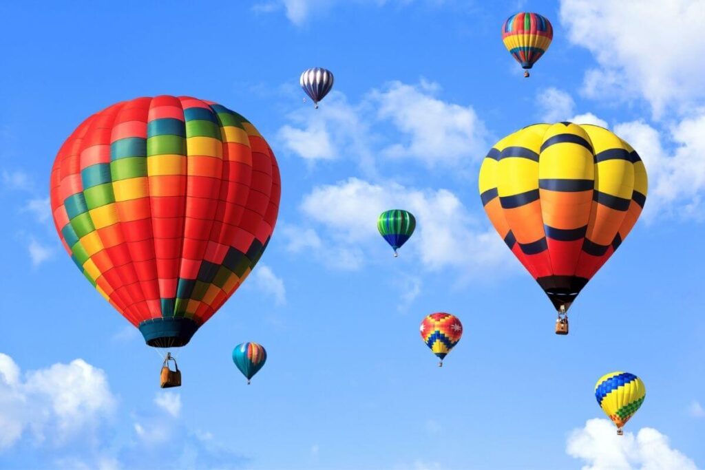 8 hot air balloons in vibrant colors of red blue yellow orange and green fly in a beautiful blue sky with a few white clouds