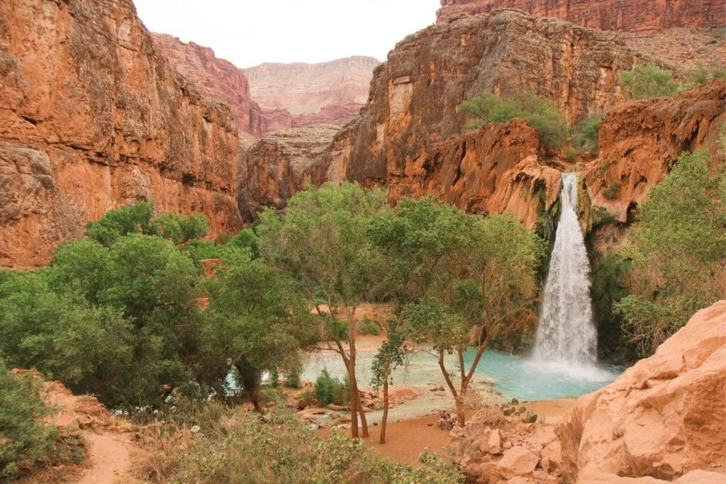 an image of Havasu Falls from a peak above the falls looking down into the red rock canyon with sparse green trees and pools of blue water