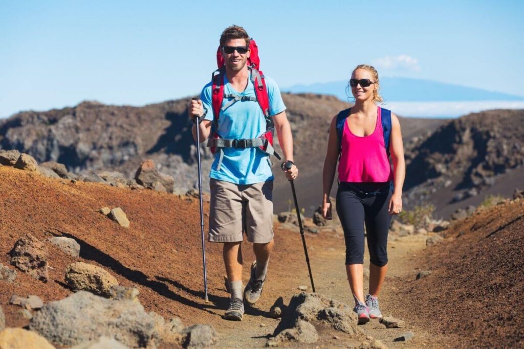 a young man and a young woman dressed in summer clothes and sunglasses wearing backpacks and carrying walking sticks hike along a dirt path in the rugged rocky mountains with a beautiful blue sky in the background