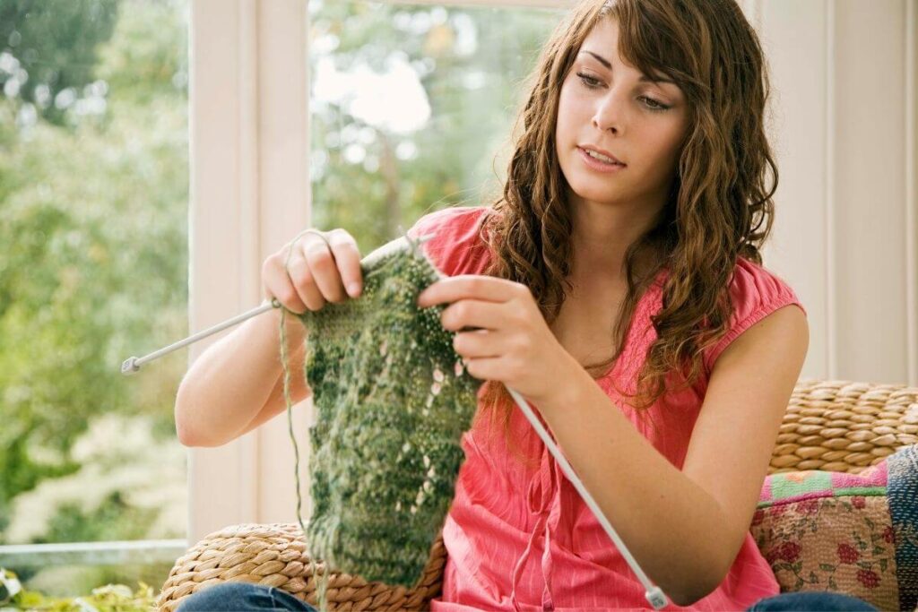 a young woman with brown curly hair learns to knit with green yarn as she sits and relaxes in front of a window