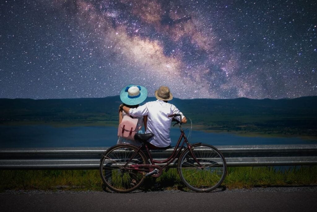 a male and female couple enjoy creative hobbies by bicycling to the edge of a lake and stargazing at the milky way in the night sky