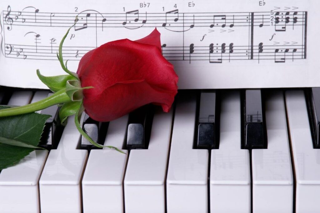 a close up image of piano keys with a sheet of music standing on the black keys and a beautiful red rose laying across the keys