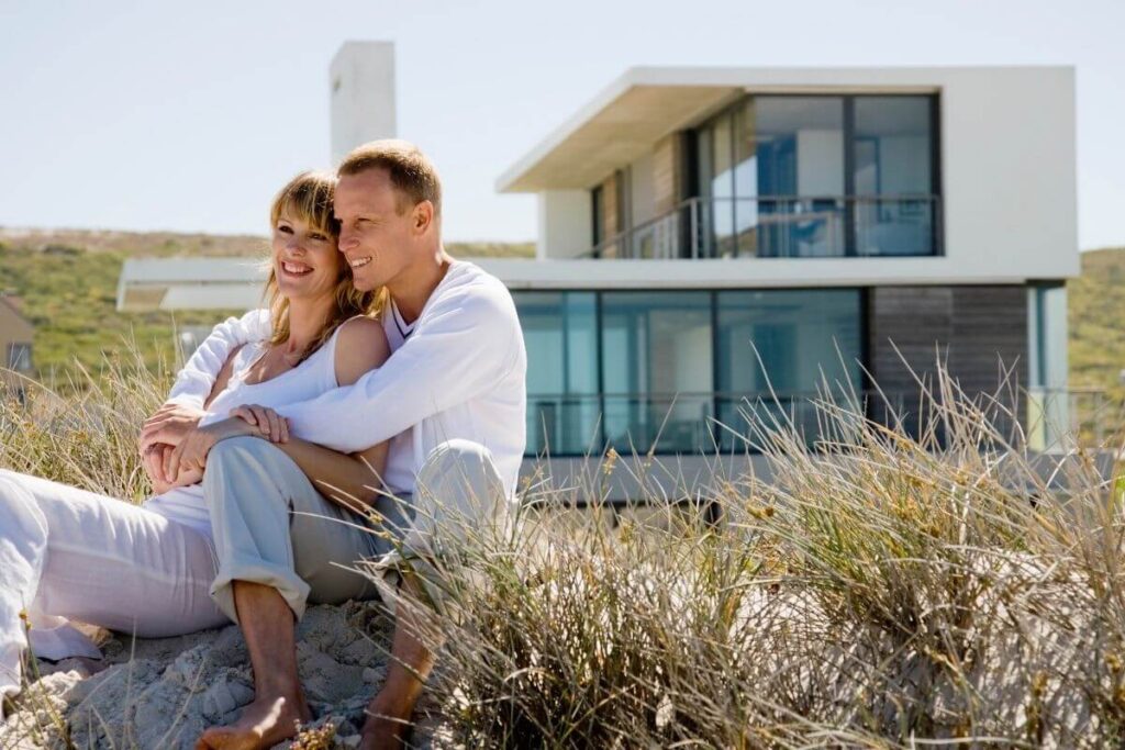 young man and woman sit together in water grasses at the beach in front of a beautiful new beach house sitting on a hill behind them