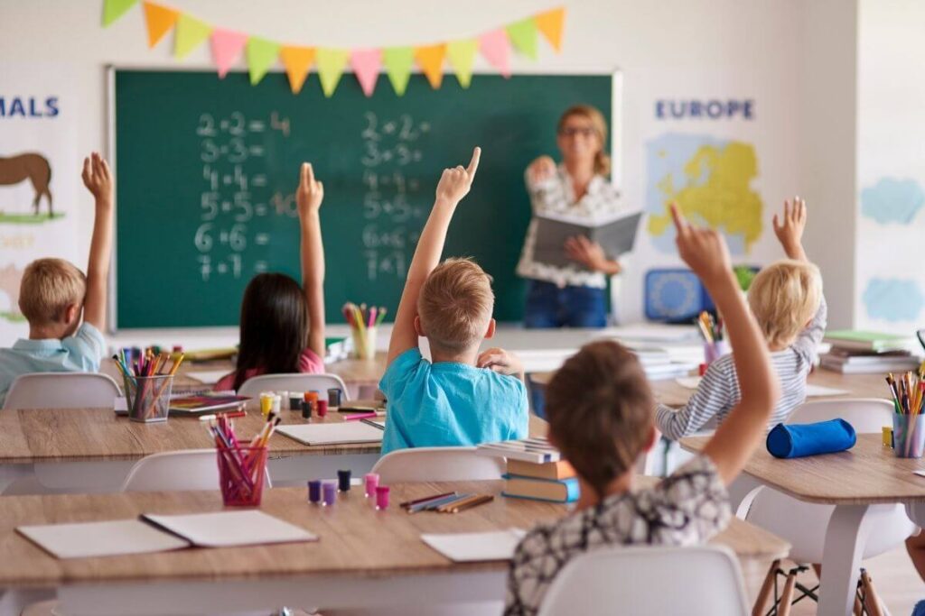 a classroom of students raise their hands to answer a question and the teacher stands in front of the classroom pointing to someone to answer