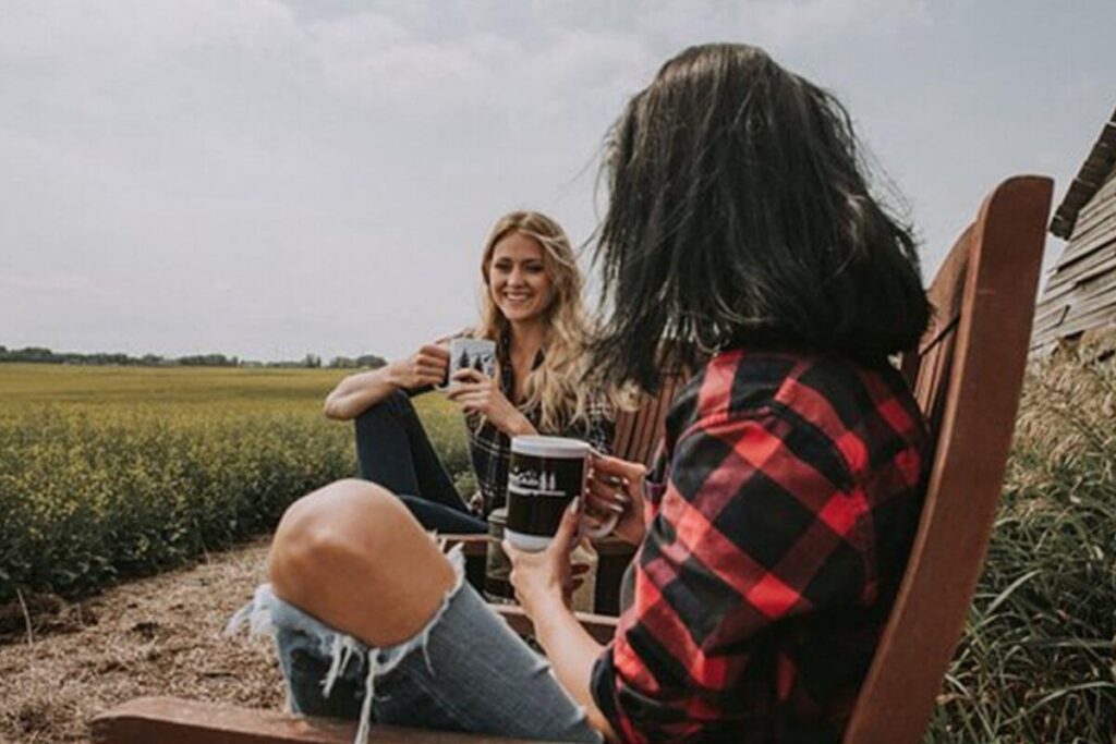 two young women dressed in jeans and flannel shirts sit in wooden chairs on the edge of a corn field talking and laughing about trusting that you can live life on your own terms and drinking hot chocolate in mugs