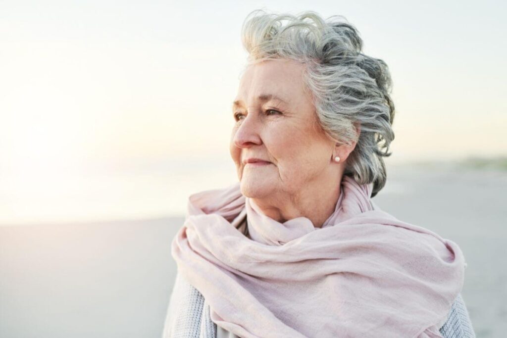 attactive elderly woman with gray hair smiles into the breeze that blows her pale pink scarf