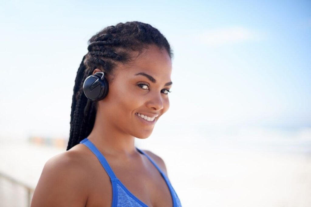 beautiful young woman with her dark hair in braids and wearing earphones smiles at a camera with a beautiful blue sky in the background because she is happy and living life on her own terms