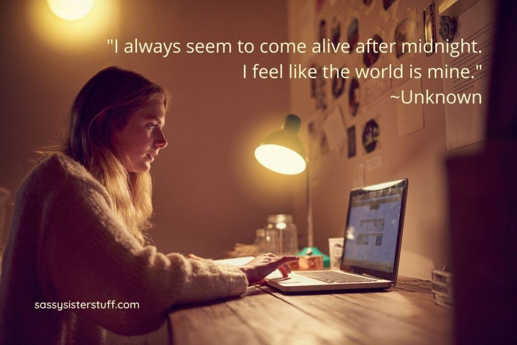 a young woman sits at her desk in a dark room with a lamp shining on her laptop as she works because she is a night owl