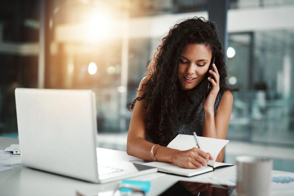 a young happy dark haired woman talks on the phone and works at her laptop in a office at night because she's a night owl