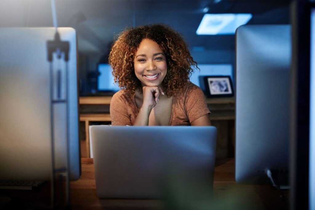 a dark skinned happy young woman sits at an opened laptop in an empty office at night because she is a night owl