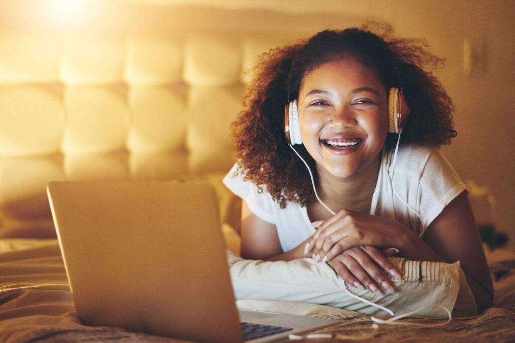 a happy teenage girl with curly hair listens to music on head phones while she works on her laptop in bed at night