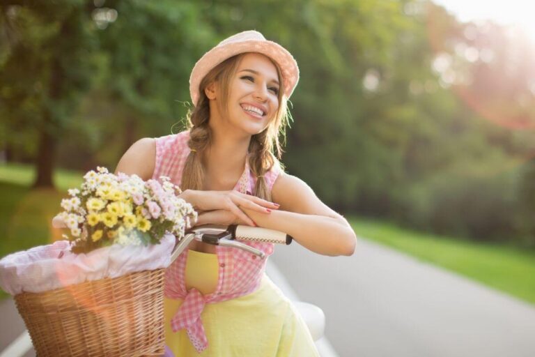 beautiful happy young woman in a pink and yellow sundress wearing a hat on a bike with a basket of pink and yellow flowers on the front