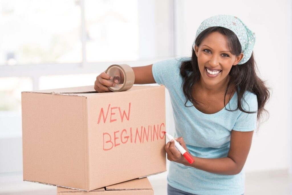 a happy young woman wearing a light teal shirt and hair scarf packs boxes and writes new beginning on the side because every journey begins with a choice and she has excitedly made a choice to move