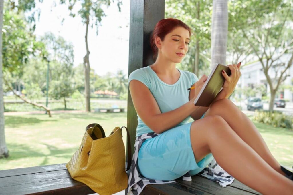 a young happy woman sits on a large window sill overlooking a beautiful green park using gratitude journals prompts to write in her journal