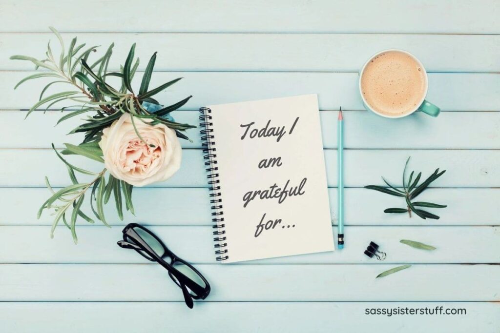 mint colored boards create a background for a cup of coffee an ivory flower with greenery black glasses a teal colored pencil and a spiral bound notebook for someone to use with gratitude journal prompts to write today I am grateful for...