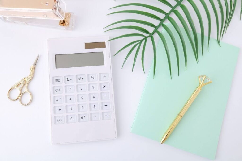 a flat lay image on a white background with a gold stapler gold scissors a white calculator a green fern leaf a gold diamond pen and a green journal