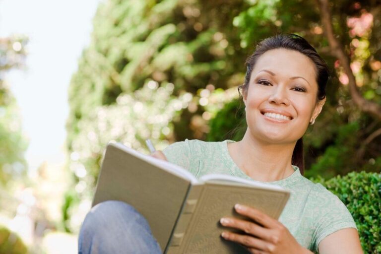 a happy woman sits outside among green trees and uses journal prompts to write in her journal