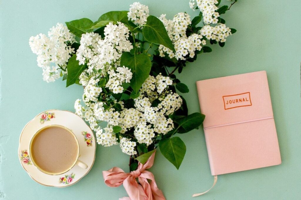 a flat lay image on a pale green background with beautiful white flowers a floral tea cup and a peach colored journal