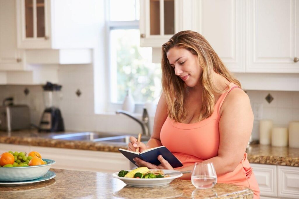 a happy woman sits on stool in her beautiful white kitchen and uses journal prompts to write in her journal while she enjoys a healthy colorful lunch