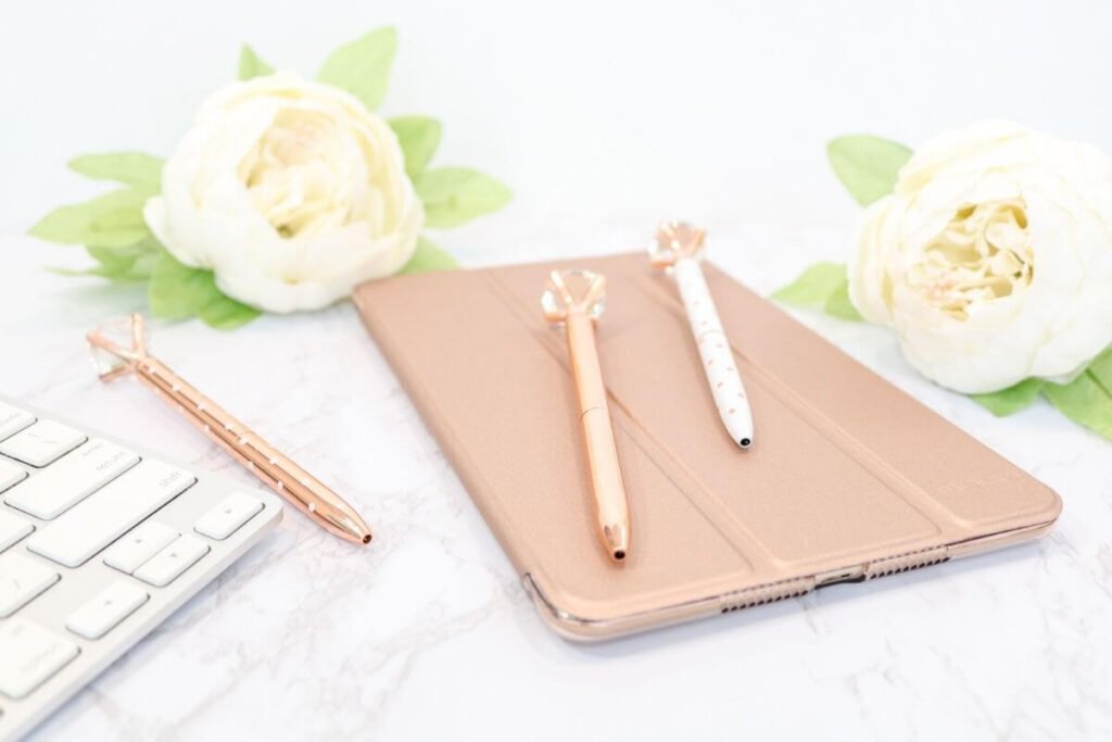 a flat lay on a white marble background with a white keyboard two white peonies three gold diamond pens and a gold tablet for journaling