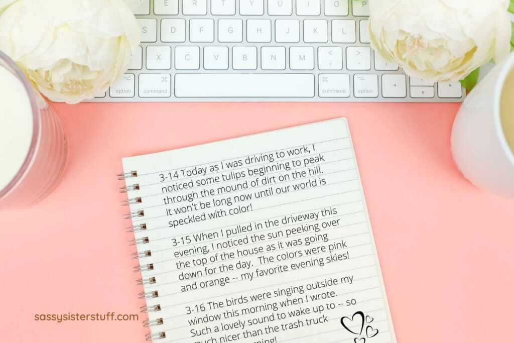 a flay lay image on a peach background with an ivory candle a white keyboard a white mug two white peonies and a spiral bound notebook with three responses to journal prompts written on a lined page