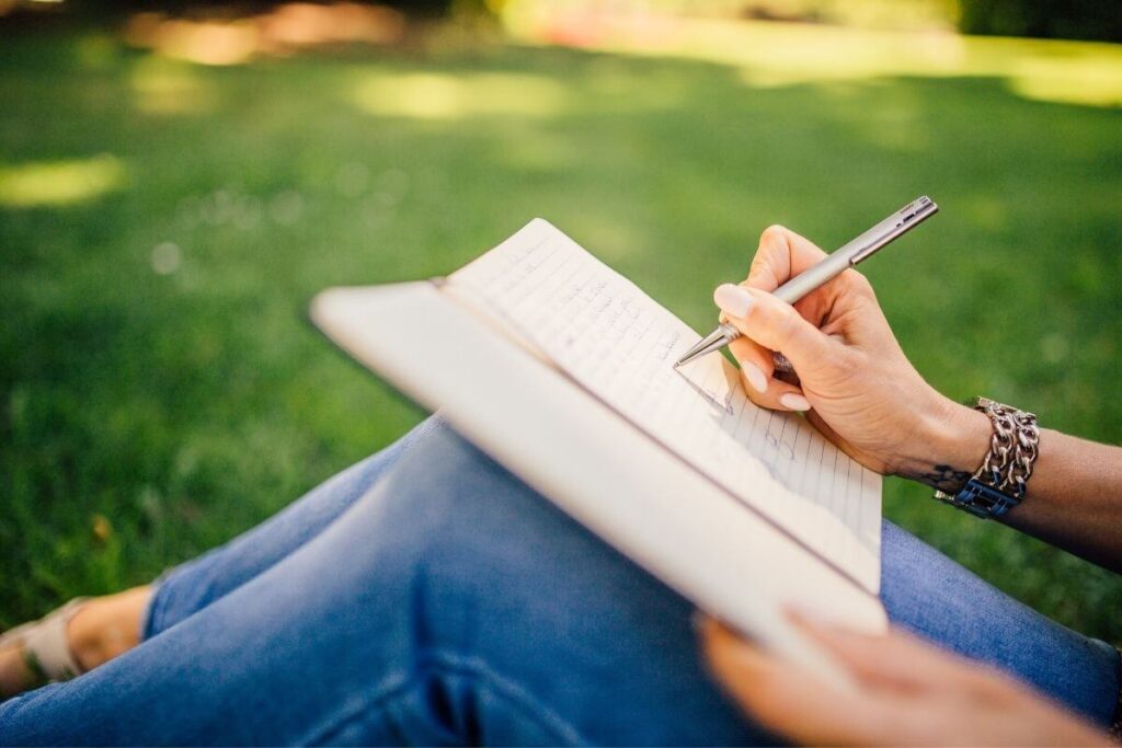 close up of a woman wearing jeans writing in a journal on her knees as she sits in a field of green grass