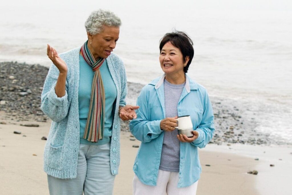 two happy middle aged women walk on the beach talking and laughing wearing blue tops and white pants as they make today a great day