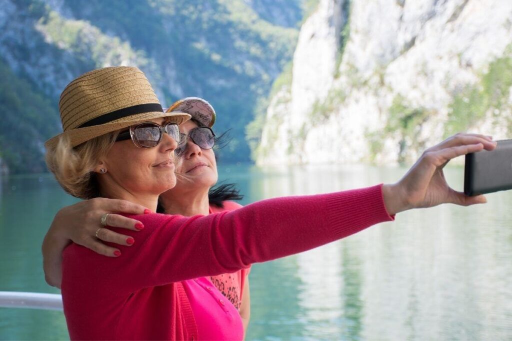 two women with hats on stand on the deck of a boat in a water passage between mountains as they take a selfie