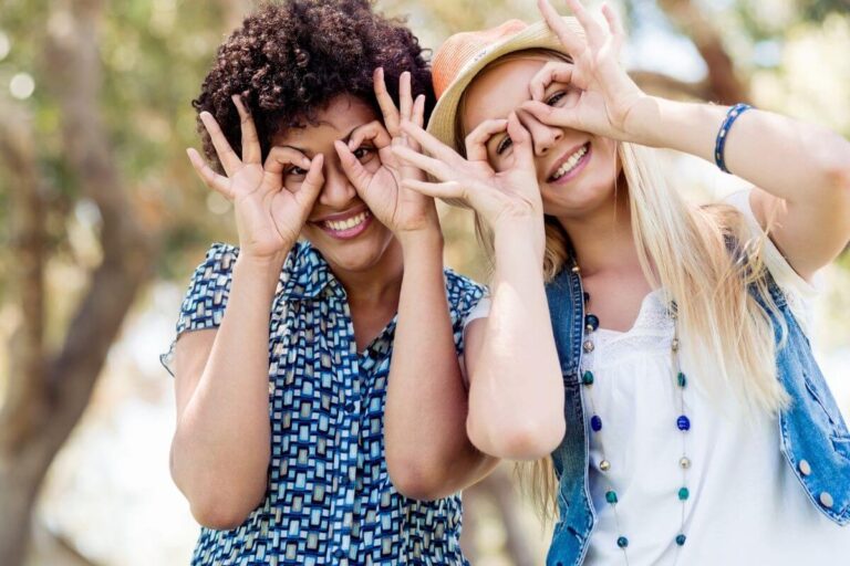 two young women wearing blue and white casual clothes laugh and make googly eyes with their pointed fingers and thumbs over their eyes on a sunny day outside in a park