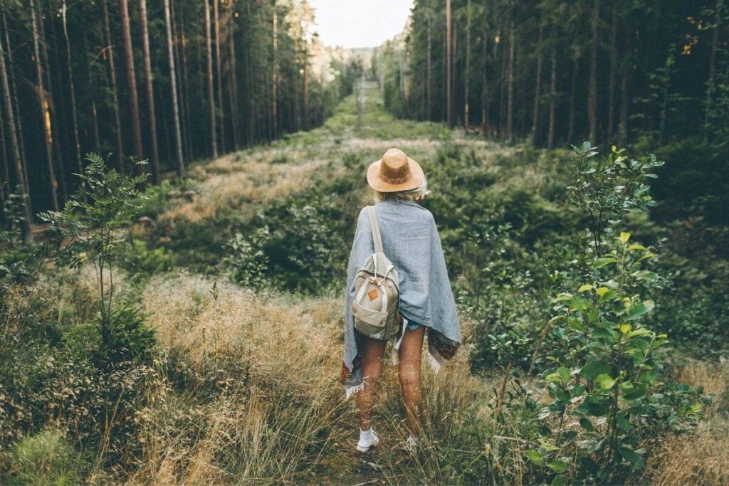 female hiker with a backpack stands in the forest looking down a long green path between tall trees