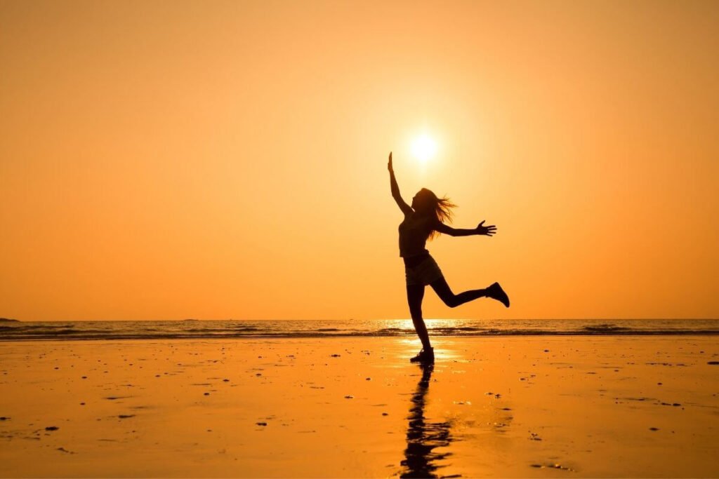 female silhouette stands on a beach in a golden sunset and kicks up one leg and both arms in celebration of the quote Do What You Can With What You Have Where You Are