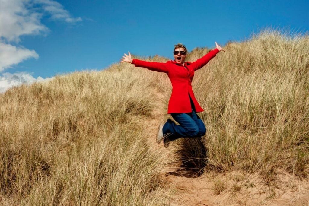 middle aged woman in jeans and a red blazer jumps with excitement in a hill of water grasses