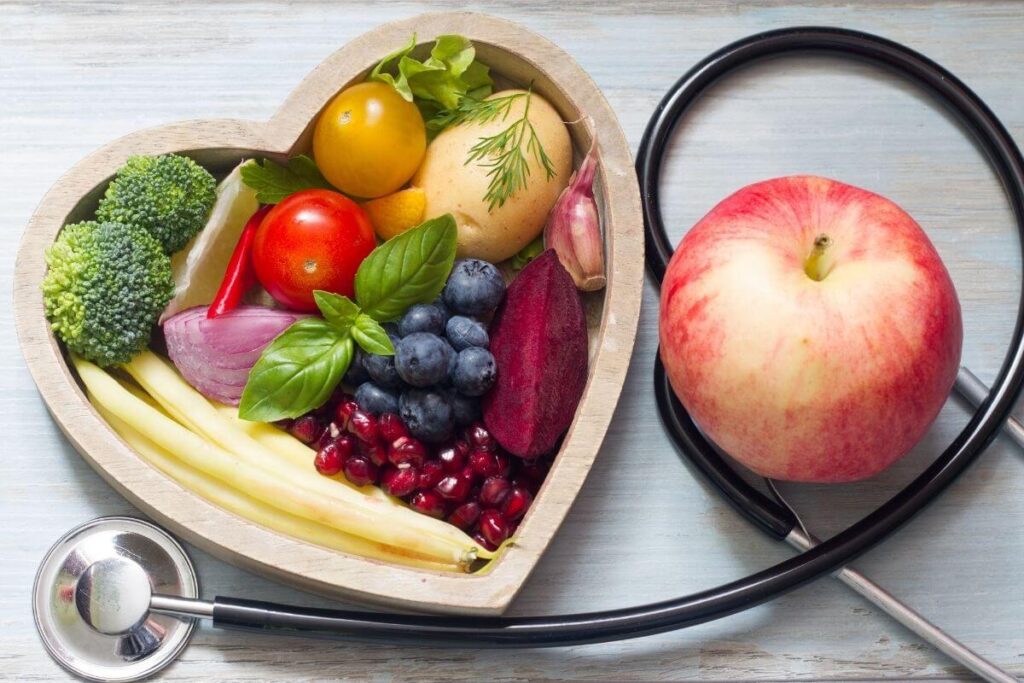 top view of a wooden heart shaped bowl with fresh fruit and vegetables in it and an apple sitting next to it with a stethoscope wrapped loosely around the apple