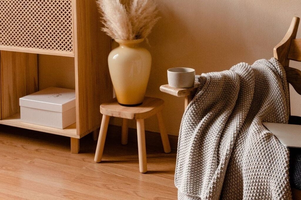 a peaceful corner with a small bookshelf a stool with a vase and fern and a chair with a mug and a blanket all in calming colors of beige and tans