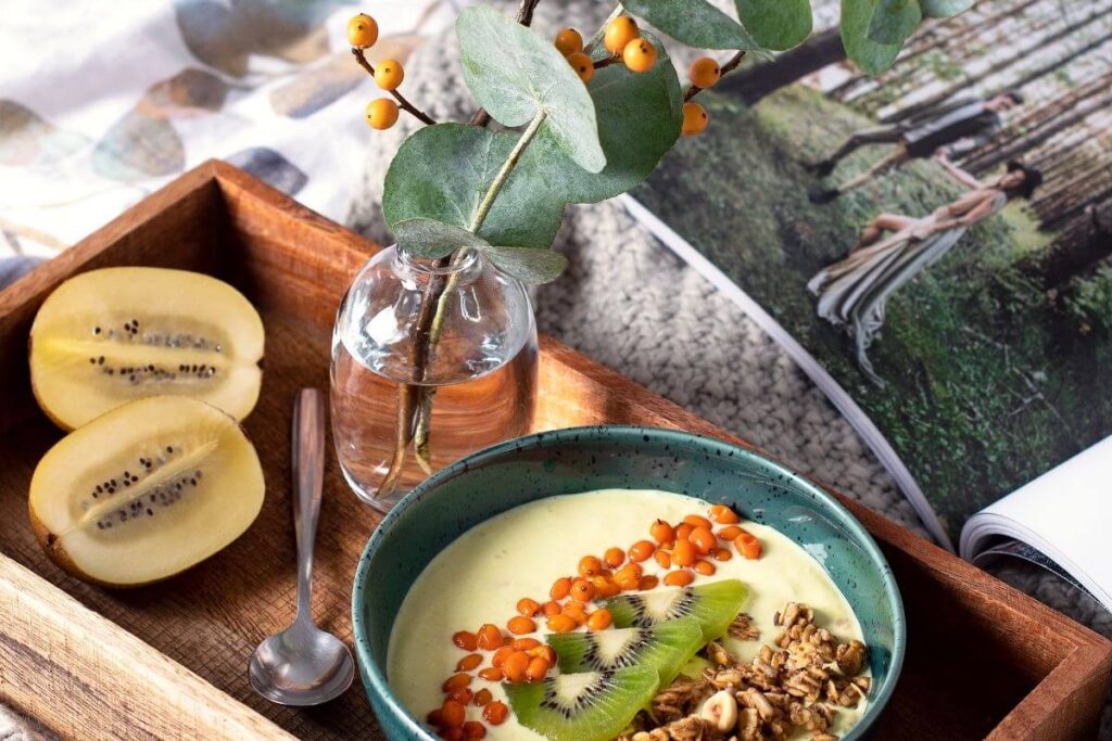 a healthy breakfast on a wooden tray with grains and oats in a bowl a kiwi cut in half and a leave and berries in a small vase of water