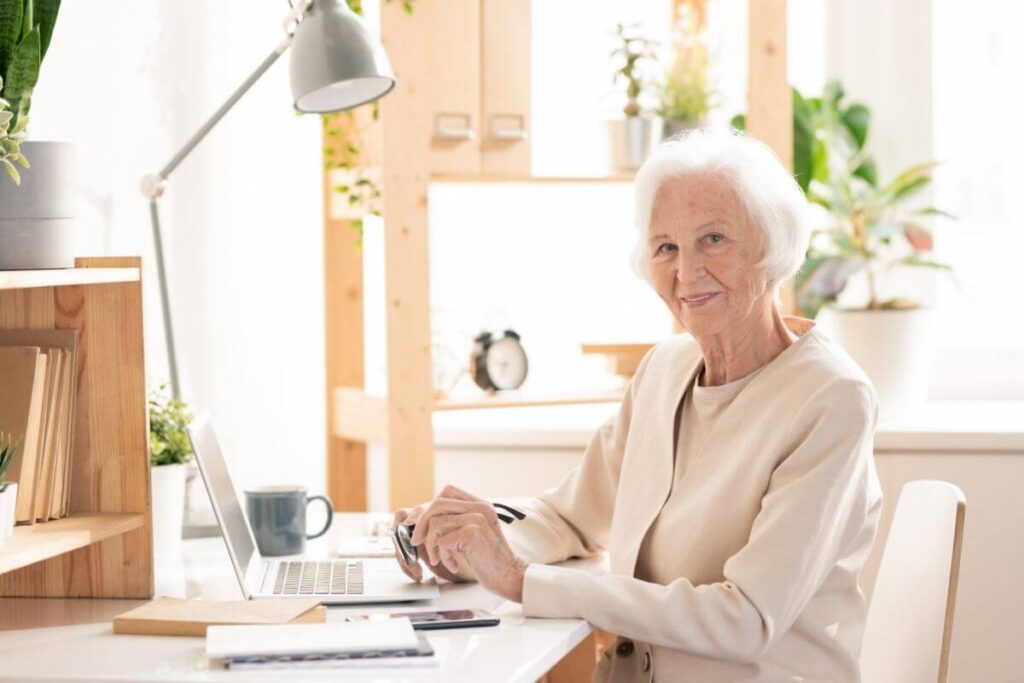 a well dressed happy senior woman sits in her home office with her journal prompts for entrepreneurs on her desk with her laptop and a light and some files for her business